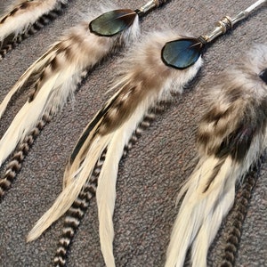 May include: Three white and brown feathers with black accents and blue accents are attached to silver cords with beads. The feathers are arranged in a row, with the longest feather at the bottom and the shortest feather at the top.