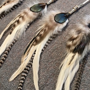 May include: Three white and brown feathers with black accents and blue accents are attached to silver cords with beads. The feathers are arranged in a row, with the longest feather at the bottom and the shortest feather at the top.