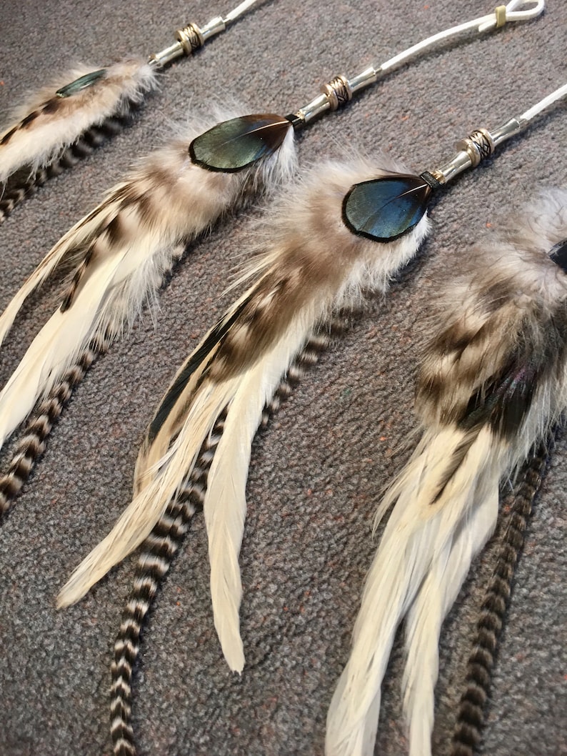 May include: Three white and brown feathers with black accents and blue accents are attached to silver cords with beads. The feathers are arranged in a row, with the longest feather at the bottom and the shortest feather at the top.