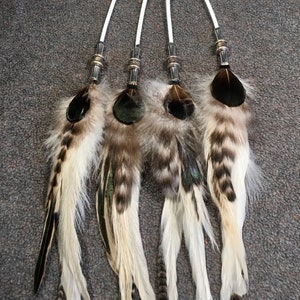 May include: Four white feather hair ties with black and brown accents. Each tie has a silver metal bead and a white string.