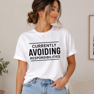 May include: A white short-sleeved t-shirt featuring the black text "CURRENTLY AVOIDING RESPONSIBILITIES" centered on the chest between two horizontal black lines. The shirt has a crew neckline and is paired with light blue denim jeans.