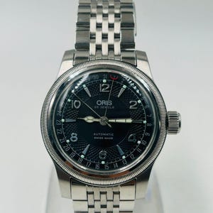 Może przedstawiać: Zegarek na rękę ze stali nierdzewnej z czarną tarczą. Tarcza zegarka zawiera markę "ORIS", tekst "AUTOMATIC SWISS MADE" i cyfrę 12. Zegarek ma srebrną bransoletę i koronkę po prawej stronie. Zegarek ma średnicę około 4 cm.