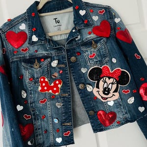 Puede incluir: Chaqueta vaquera azul adornada con corazones de lentejuelas rojas, corazones blancos y pedrería. Presenta un parche de Minnie Mouse y un lazo rojo. La chaqueta es talla 14.