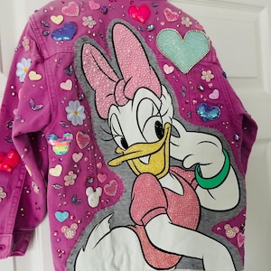 May include: A lilac denim jacket decorated with sequined hearts, flowers, and a large Daisy Duck graphic. The jacket has a button-down front and is embellished with rhinestones. The Daisy Duck design is outlined in grey and has a pink dress and bow.