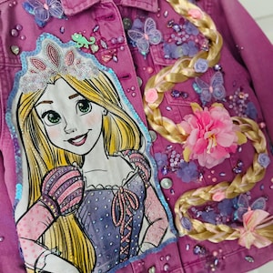 May include: A magenta denim jacket decorated with a princess cartoon illustration, sequins, and butterflies. A braided hair design is on the right side. The jacket has a button-up front and is a vibrant shade of pink.