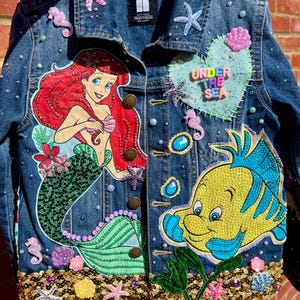May include: A blue denim jacket decorated with an appliqué of a red-haired mermaid and a yellow fish. The jacket features various sea-themed embellishments including starfish, seahorses, shells, pearls, and a heart patch with the text UNDER THE SEA.