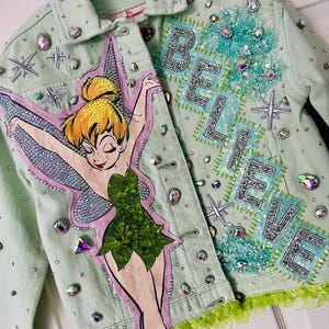 May include: A pale green denim jacket decorated with a Tinkerbell illustration, rhinestones, and the word "BELIEVE" in silver sequins. The jacket has a green sequined trim and various embellishments.