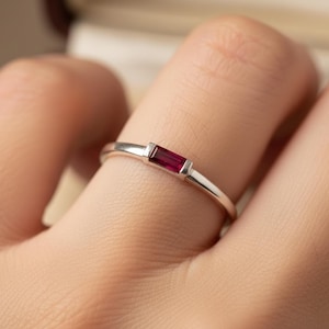 Puede incluir: Un anillo de plata con una gema rectangular de color rojo intenso. La gema está engastada horizontalmente en un diseño minimalista. El anillo se muestra en un dedo, destacando la elegante simplicidad de la joya.