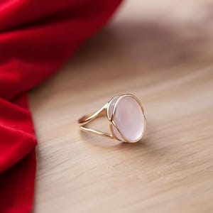 Könnte beinhalten: Ein goldener Ring mit einem großen, ovalen, hellrosa Edelstein. Der Ring hat ein zartes, offenes Design mit einem geteilten Band. Der Edelstein ist in einer weißen Lünette gefasst. Der Ring wird auf einer hellen Holzoberfläche mit rotem Stoff im Hintergrund präsentiert.