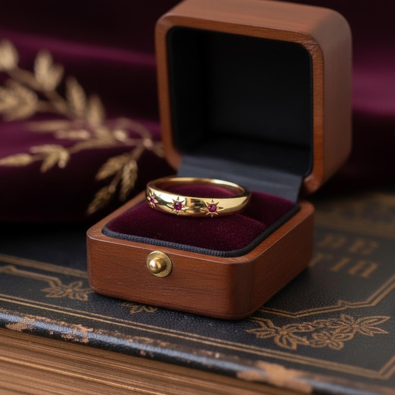 May include: A gold ring with three red star-shaped accents, presented in an open wooden ring box with a burgundy velvet lining. The box is displayed on a dark surface. The ring is the focal point.