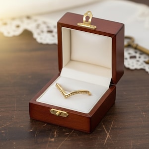 Peut inclure: Une bague dorée en forme de V avec de petits ornements, présentée dans un écrin à bijoux en bois foncé ouvert avec un intérieur blanc. La boîte a un fermoir doré et est posée sur une surface en bois foncé.