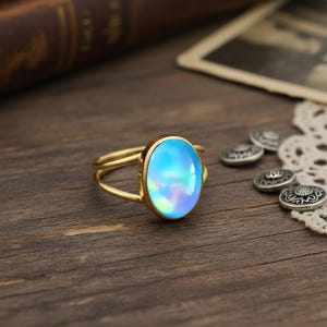 May include: A gold ring with a large, oval, iridescent blue gemstone. The ring has a double band design. The gemstone reflects shades of blue, purple, and green. The ring is set on a wooden surface.