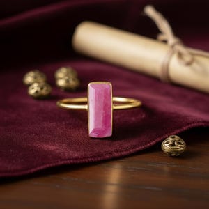 Könnte beinhalten: Ein goldener Ring mit einem rechteckigen, rosa Edelstein. Der Ring ist vor einem dunkelroten Samthintergrund platziert, mit einer Schriftrolle und dekorativen Perlen im Hintergrund. Das Design des Rings ist schlicht und elegant.