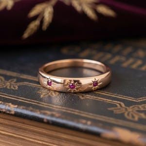 May include: A gold ring with three starburst designs, each set with a small, round, red gemstone. The ring's polished surface is set against a dark background, highlighting its intricate details. A piece of jewellery.