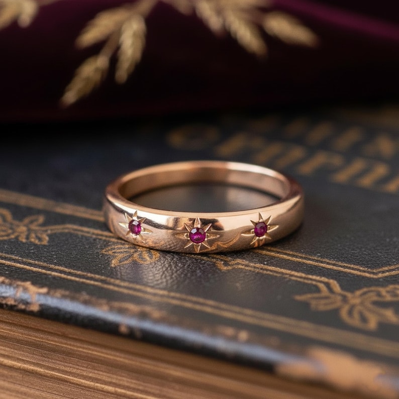 May include: A gold ring with three starburst designs, each set with a small, round, red gemstone. The ring's polished surface is set against a dark background, highlighting its intricate details. A piece of jewellery.
