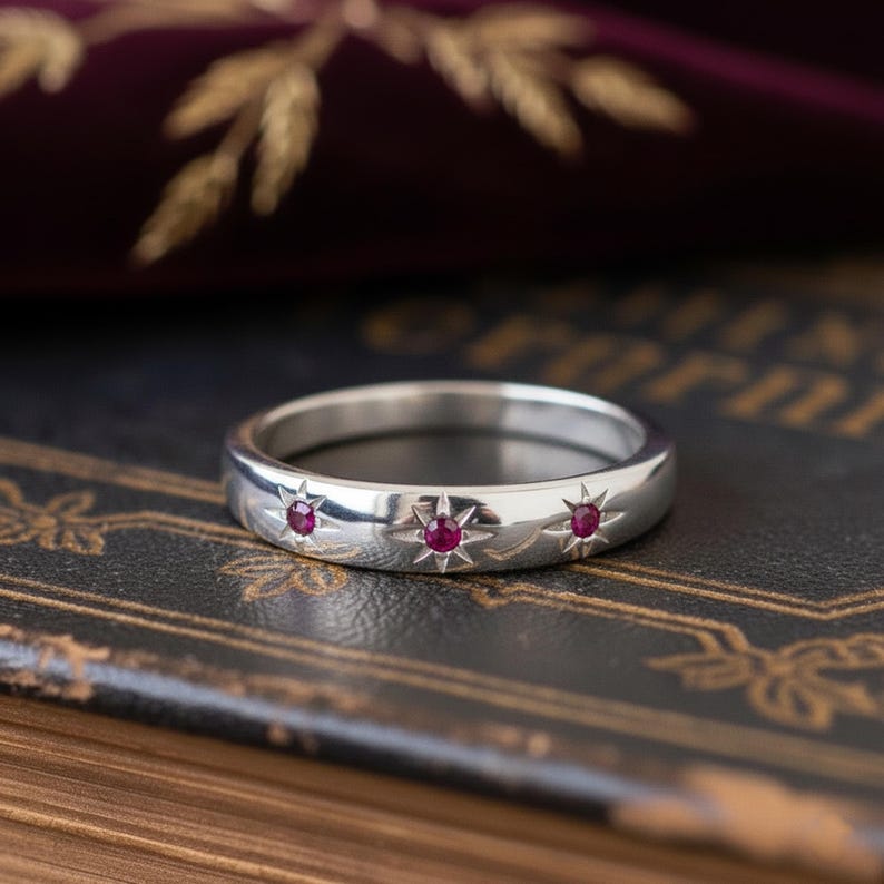 May include: A polished silver ring, adorned with three starburst motifs, each set with a small, round, red gemstone. The ring's design is set against a dark, textured backdrop. The ring is a piece of jewellery.