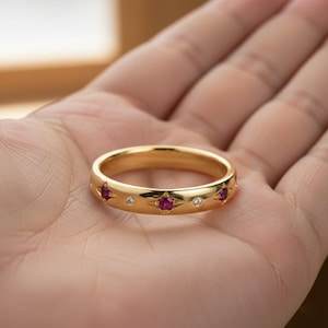 Puede incluir: Un anillo dorado con piedras rojas engastadas en diseños de estrella, alternando con pequeñas piedras transparentes. El anillo se muestra en una mano, resaltando su superficie pulida y sus detalles intrincados. Una pieza de joyería clásica.