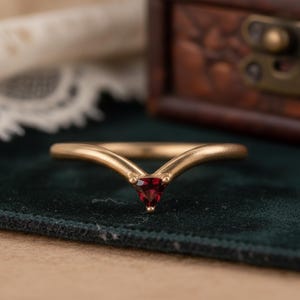 Può includere: Un anello dorato con un design a forma di V, con una gemma triangolare rosso scuro al centro. L'anello è posto su una superficie di velluto verde scuro, con uno sfondo sfocato che include una scatola di legno.