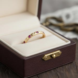 Può includere: Un anello d'oro con piccole gemme rosse incastonate nella fascia, presentato in un portagioie marrone con interno color crema. L'anello presenta un motivo a stella inciso lungo la fascia.