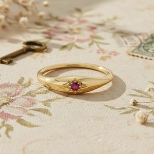 Puede incluir: Un anillo de oro con un diseño de estrella y una gema roja central. El anillo tiene un acabado liso y pulido y un estilo clásico y elegante. El anillo es de talla 14.