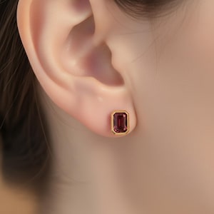 Könnte beinhalten: Ein goldener Ohrstecker mit einem rechteckigen, tiefroten Edelstein. Der Edelstein ist in eine goldene Lünette gefasst, was einen klassischen und eleganten Look erzeugt. Der Ohrring wird an einem Ohr gezeigt.