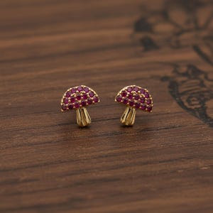 Puede incluir: Un par de pendientes de botón con forma de seta dorada. Las tapas de las setas están cubiertas de pequeñas piedras preciosas rojas y redondas. Los pendientes se exhiben sobre una superficie de madera marrón oscuro.