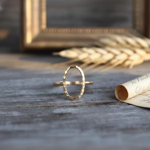 May include: A gold-colored ring with an oval-shaped hammered design. The ring is set against a blurred background with a gold frame, wheat stalks, and a rolled-up paper. The ring's band is thin and delicate.