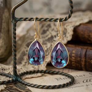 May include: A pair of gold-colored drop earrings with teardrop-shaped, faceted gemstones. The gemstones have a color gradient of deep purple to teal blue. The earrings are suspended from a decorative, dark metal display stand.