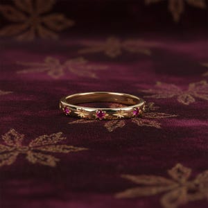 Puede incluir: Un anillo de oro con diseños de estrellas y pequeñas gemas rojas redondas. El anillo se encuentra sobre un fondo rojo oscuro con un patrón floral.