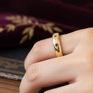 May include: A gold ring featuring two small, round, red gemstones. The ring has a smooth, polished surface and is set with the gemstones in a starburst design. The ring is on a finger.