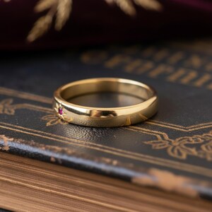 May include: A gold ring featuring a small red gemstone and etched sunburst pattern. The ring is displayed on a dark surface, emphasizing its polished finish and delicate design. A timeless piece of jewellery.