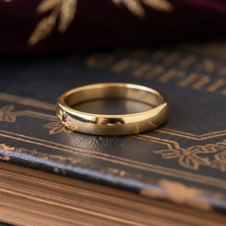May include: A gold ring featuring a small red gemstone and etched sunburst pattern. The ring is displayed on a dark surface, emphasizing its polished finish and delicate design. A timeless piece of jewellery.