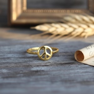 May include: A gold ring with a peace symbol design. The ring is displayed on a wooden surface, with a blurred background including a picture frame and wheat stalks. The ring's design is the main focus of the image.