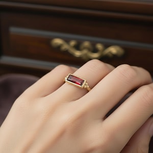 Puede incluir: Un anillo de oro con una piedra preciosa rectangular de color rojo intenso. El anillo tiene una banda simple con pequeñas cuentas doradas decorativas a cada lado de la piedra preciosa. El anillo se muestra en un dedo.