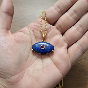 Puede incluir: Un collar de cadena dorada con un colgante ovalado de piedra azul oscuro. El colgante tiene una pequeña gema roja redonda en el centro y está adornado con puntas doradas. El collar se muestra en una mano.