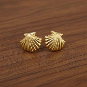 May include: A pair of gold-colored seashell stud earrings. The earrings are small, with a detailed, fan-like design. The earrings are displayed on a wooden surface.