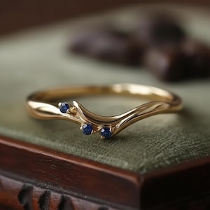 Op de afbeelding: Een goudkleurige ring met een gebogen ontwerp, met drie kleine, ronde, blauwe edelstenen. De ring is geplaatst tegen een zachte groene achtergrond, met een wazig houten element op de achtergrond. Het ontwerp van de ring is elegant en delicaat.