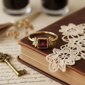 May include: A gold ring with a square, deep red gemstone. The ring has leaf-like designs on either side of the gemstone. The ring is displayed on a brown book with a vintage key and lace doily.