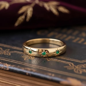 May include: A gold ring featuring three emerald-green gemstones set within starburst patterns. The ring rests on a dark, textured surface, with a blurred background showing gold leaf details.