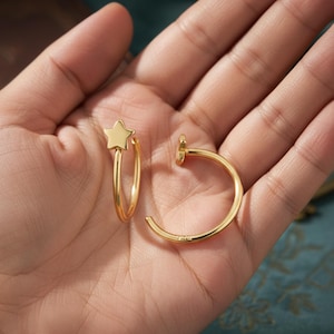 Puede incluir: Un par de pendientes de aro dorados, uno con un dije de estrella. Los pendientes se muestran en una mano. Los pendientes tienen un diseño sencillo y elegante. Están hechos de metal dorado.