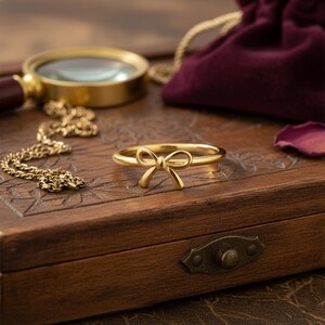 May include: A gold bow ring, set on a wooden box with a magnifying glass and a gold chain. The ring's bow design is the central feature, adding a touch of elegance. Other items include a velvet pouch and a rose petal.