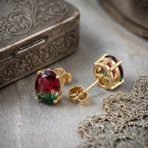 Puede incluir: Un par de pendientes de oro con piedras preciosas de corte ovalado. Las piedras muestran un degradado de colores, incluyendo rojo, verde y un toque de amarillo. Los pendientes están sobre un fondo gris suave, con una caja plateada decorativa en el fondo.