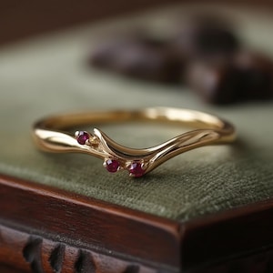 Op de afbeelding: Een goudkleurige ring met een gebogen ontwerp, met drie kleine, ronde, rode edelstenen. De ring heeft een delicate, organische vorm, die doet denken aan een bloemen- of natuurlijk thema.