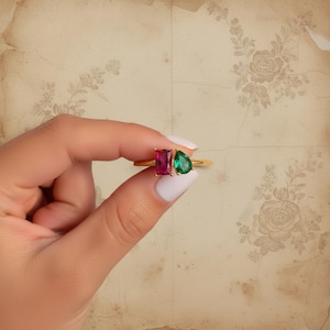 Può includere: Un anello d'oro con una gemma rosa rettangolare e una gemma verde a forma di pera. L'anello è esposto su uno sfondo floreale vintage.
