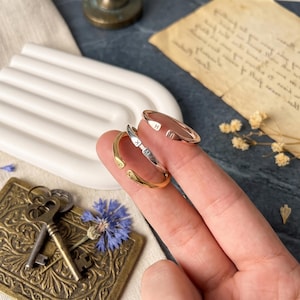 May include: Three open-ended rings in gold, silver, and rose gold, each with engraved initials. The rings are displayed on a finger, showcasing their minimalist design. Other items in the background include keys, a blue flower, and a vintage paper.