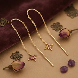 Op de afbeelding: Gouden oorhangers met ster-vormige bedels. Elke ster is versierd met kleine, roze edelstenen. De oorbellen worden gepresenteerd op een beige oppervlak met gedroogde bloemen en decoratieve metalen accenten.