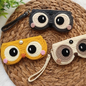 May include: Three rectangular crochet pouches featuring owl-like designs with large circular eyes, black pupils, and small pink cheeks. Each pouch includes a matching wrist strap and a central snap button closure, available in dark grey, yellow, and cream.