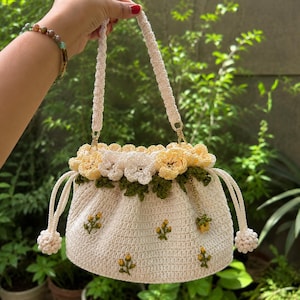 May include: A cream-coloured crochet drawstring bag featuring a top border of yellow and white crocheted flowers with green leaves. The body of the bag is decorated with small embroidered yellow flower sprigs and has a single braided handle.