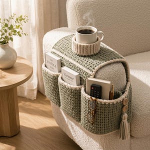 Peut inclure: Un organisateur d'accoudoir en crochet vert sauge avec plusieurs poches de rangement, un porte-gobelet intégré avec une tasse blanche, et deux boutons en bois avec un pompon. Les poches contiennent des livres, une télécommande et des lunettes.
