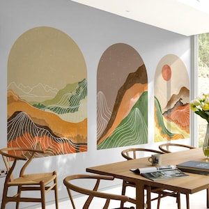 May include: Three wall art pieces depicting abstract mountain landscapes within arched frames. The artwork uses earthy tones of orange, brown, and green, with white contour lines. One piece includes a red sun. The art is displayed in a dining room setting.